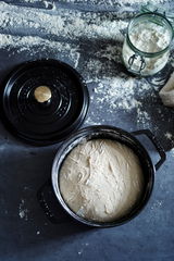 Runde schwarze Cocotte mit Teig, Deckel daneben, von oben fotografiert, auf bemehlter Oberfläche.