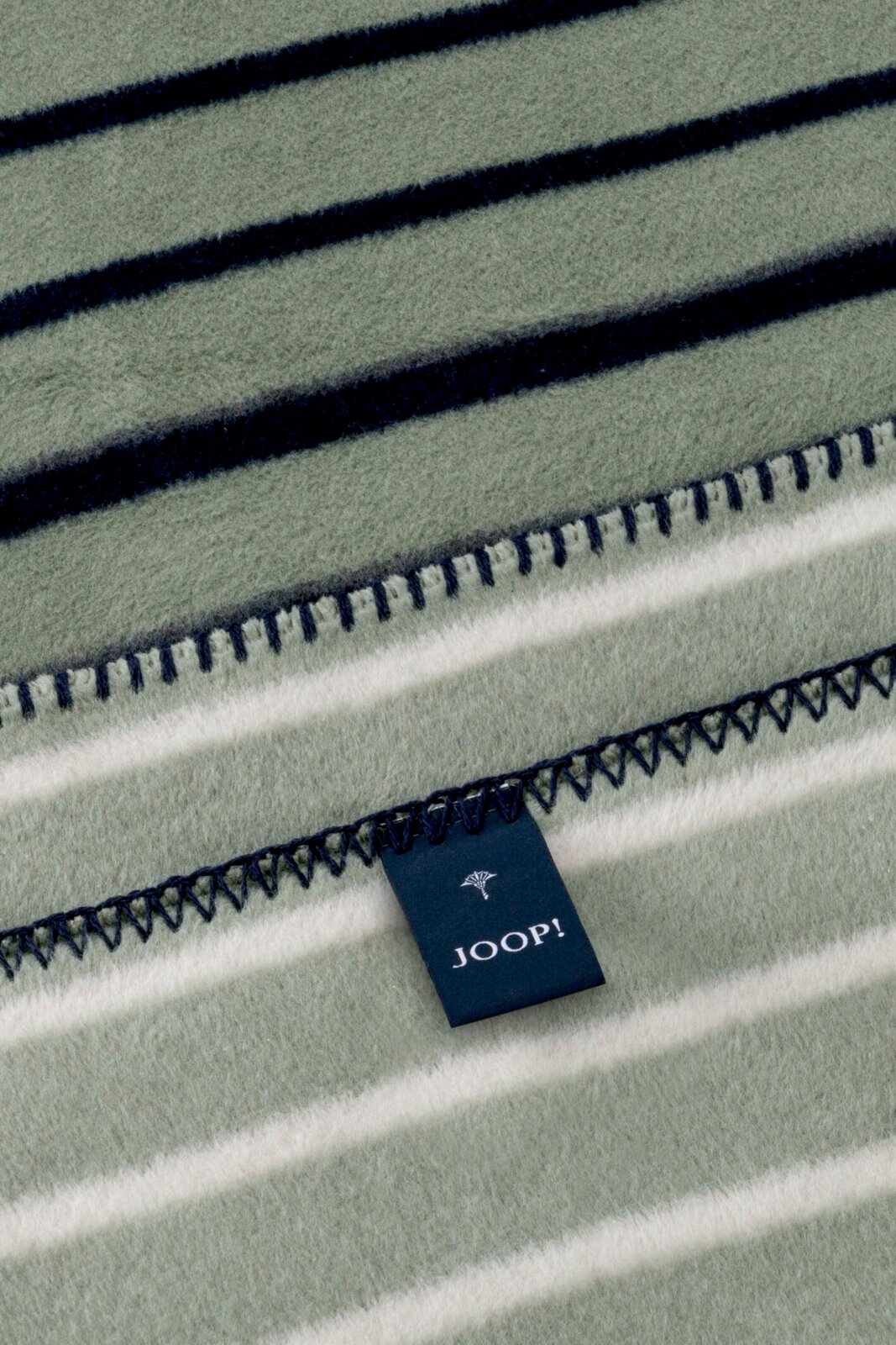 JOOP! Wohndecke 150 x 200 cm Detailaufnahme aus schräger Draufsicht: gestreifte JOOP! Wohndecke in Grau/Weiß mit dunkler Ziernaht und Markenlabel