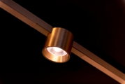 Schuine onderaanzicht close-up van een bronzen/cognackleurige cilindrische railspot met ingeschakelde LED op een rail tegen een donkere achtergrond