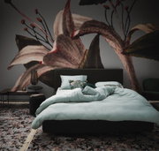 Bett mit hellblauer Bettwäsche in einem stilvoll eingerichteten Raum mit großem Blumenmuster an der Wand, Perspektive von vorne.