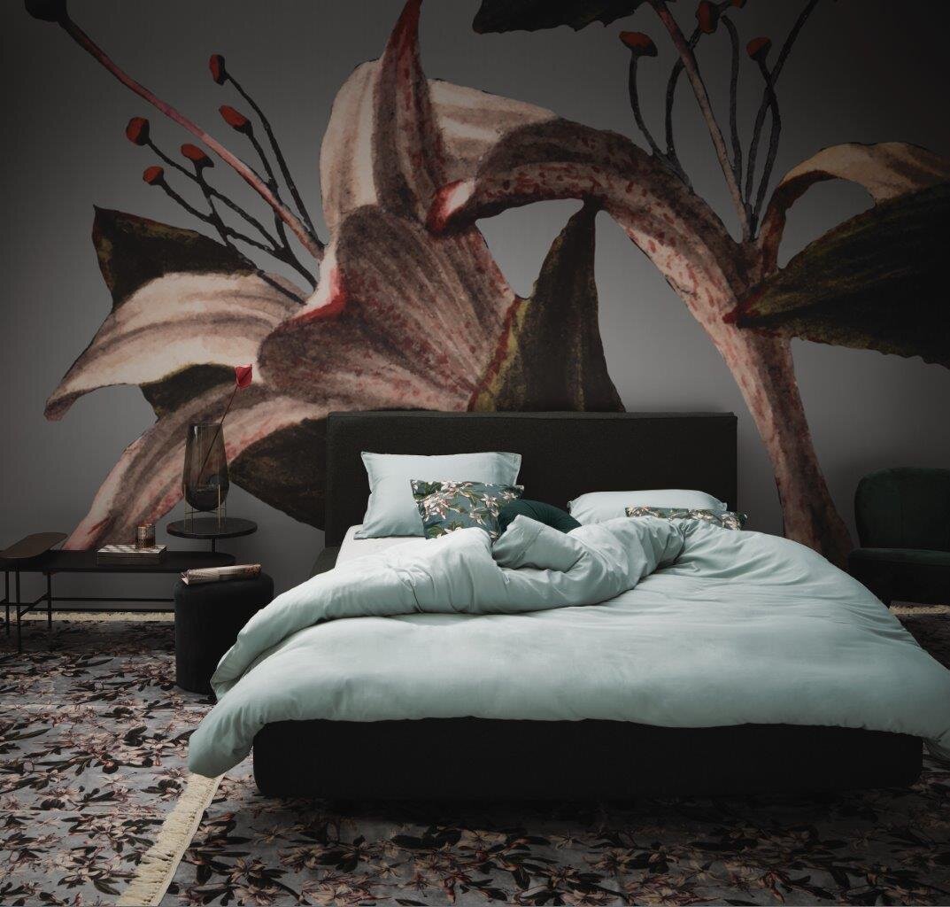 Bett mit hellblauer Bettwäsche in einem stilvoll eingerichteten Raum mit großem Blumenmuster an der Wand, Perspektive von vorne.