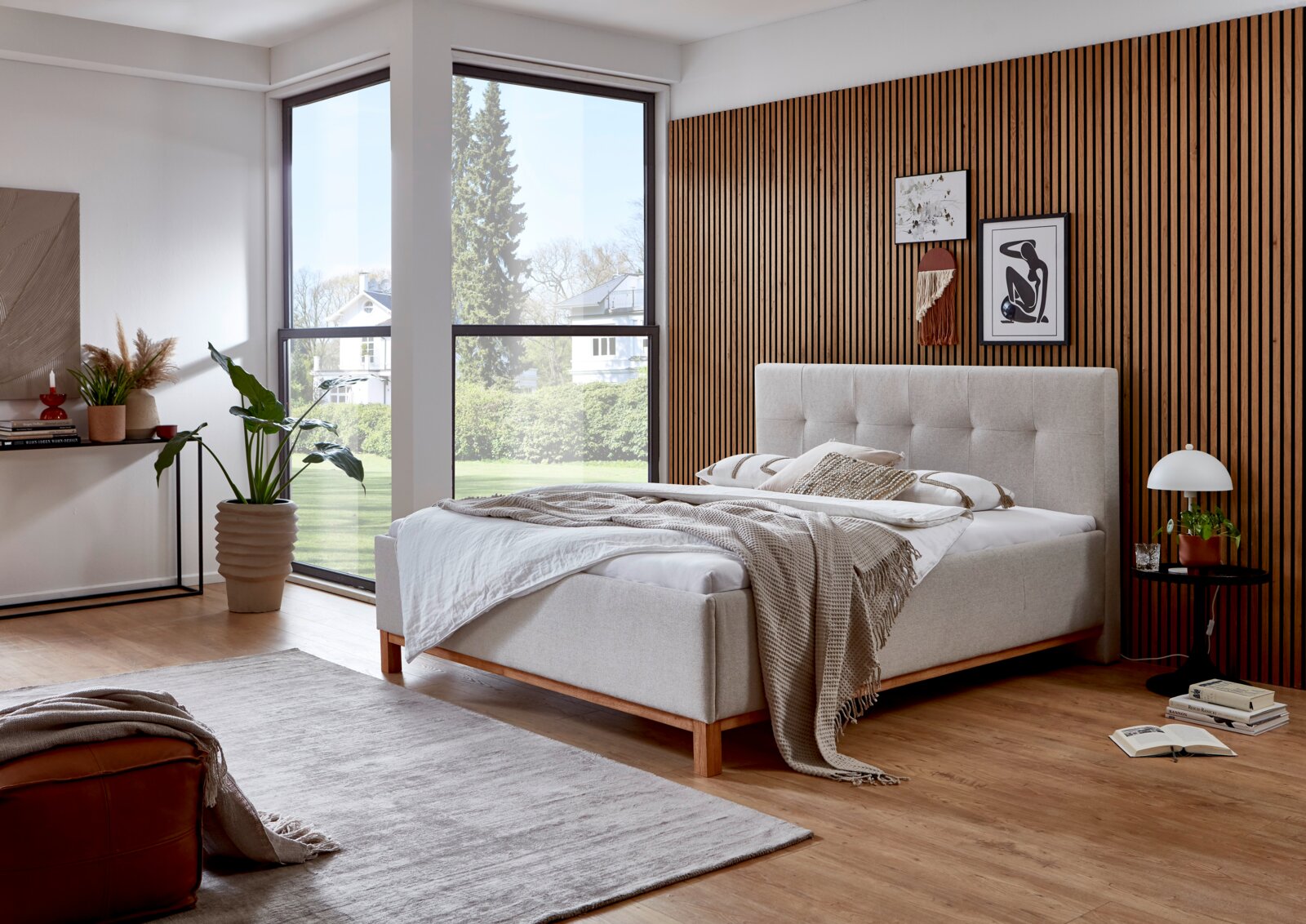Modern gestoffeerd bed in een stijlvolle slaapkamer met houten lambrisering en grote ramen, zijaanzicht.