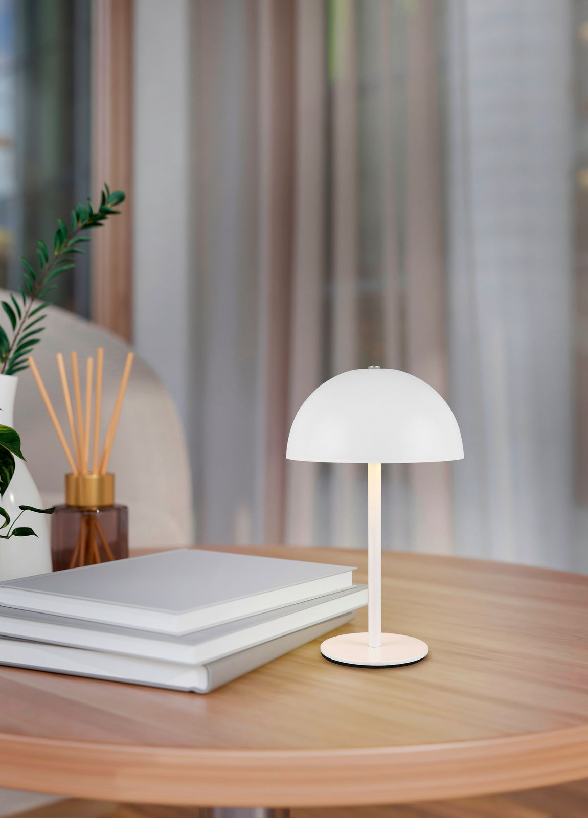 Reality Leuchten Akku-Tischleuchte MUNOZ Weiße Akku-Tischleuchte MUNOZ auf einem Holztisch, seitliche Perspektive, umgeben von Büchern und Dekoration.