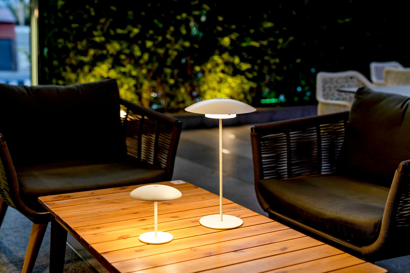 Zwei moderne, weiße LED-Tischlampen mit pilzförmigem Schirm auf einem Holz-Loungetisch zwischen zwei Outdoor-Sofas, aufgenommen aus leicht erhöhter, frontaler Perspektive bei Abendstimmung.