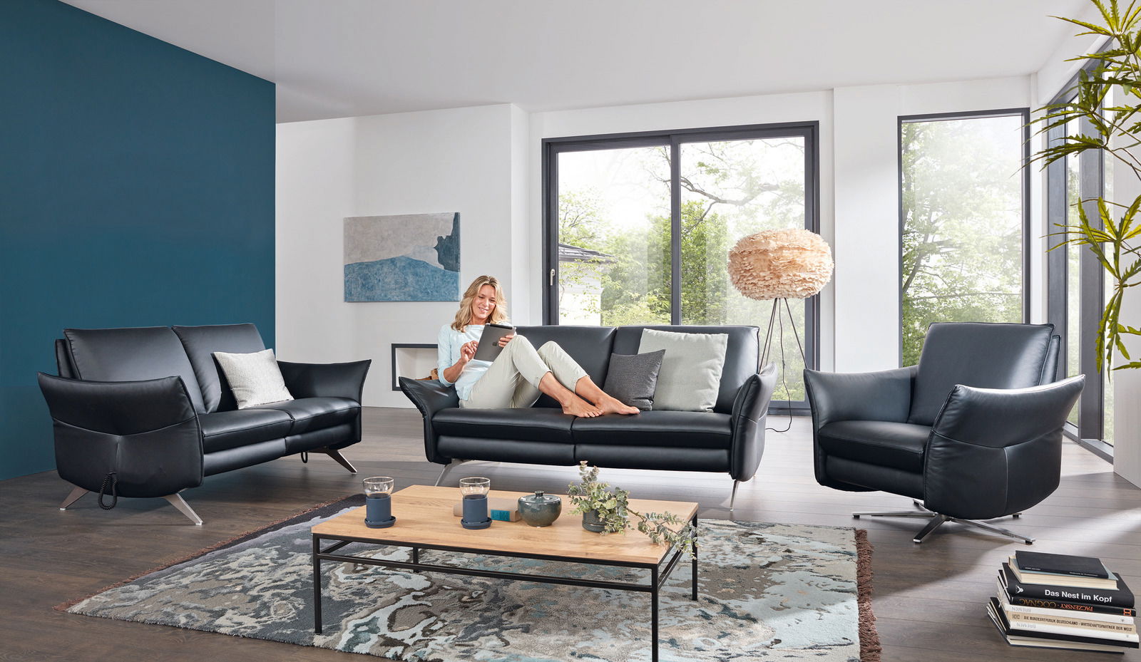 Wohnzimmer-Set mit zwei schwarzen Sofas und einem Sessel, frontal betrachtet, mit einer Frau, die auf einem Sofa sitzt und ein Tablet benutzt.