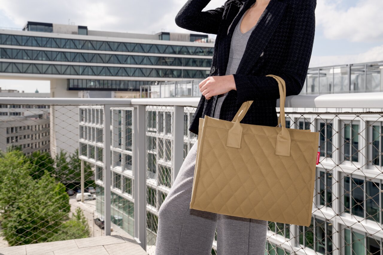 Frau auf einem Balkon mit einer beigefarbenen, rautenförmig gesteppten Einkaufstasche, seitliche Perspektive