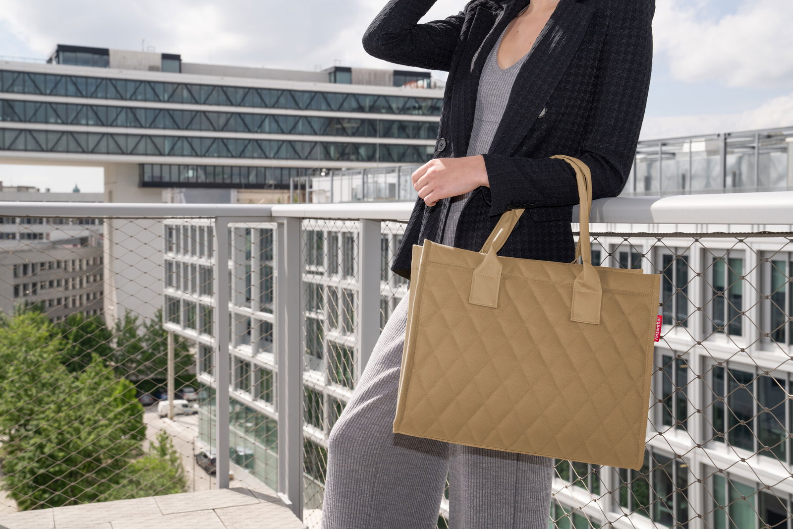 Frau auf einem Balkon mit einer beigefarbenen, rautenförmig gesteppten Einkaufstasche, seitliche Perspektive