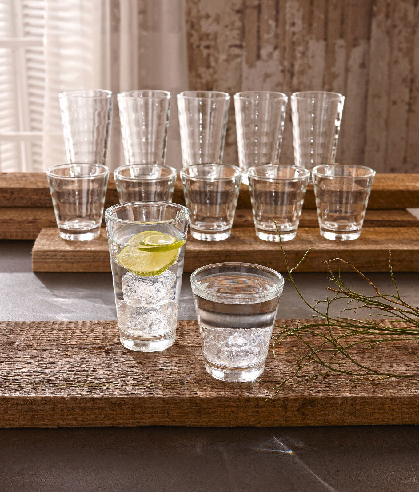 Becherset Wave 12-tlg. aus Glas, bestehend aus verschiedenen Größen, auf rustikalen Holzplatten präsentiert, mit Eiswürfeln und Limettenscheibe in einem Glas, seitliche Perspektive.