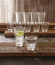 Becherset Wave 12-tlg. aus Glas, bestehend aus verschiedenen Größen, auf rustikalen Holzplatten präsentiert, mit Eiswürfeln und Limettenscheibe in einem Glas, seitliche Perspektive.