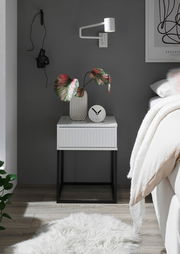 Moderne Nachtkommode mit weißer Oberfläche und schwarzen Metallbeinen, dekoriert mit einer Vase und einer Uhr, seitliche Perspektive.