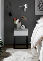 Moderne Nachtkommode mit weißer Oberfläche und schwarzen Metallbeinen, dekoriert mit einer Vase und einer Uhr, seitliche Perspektive.