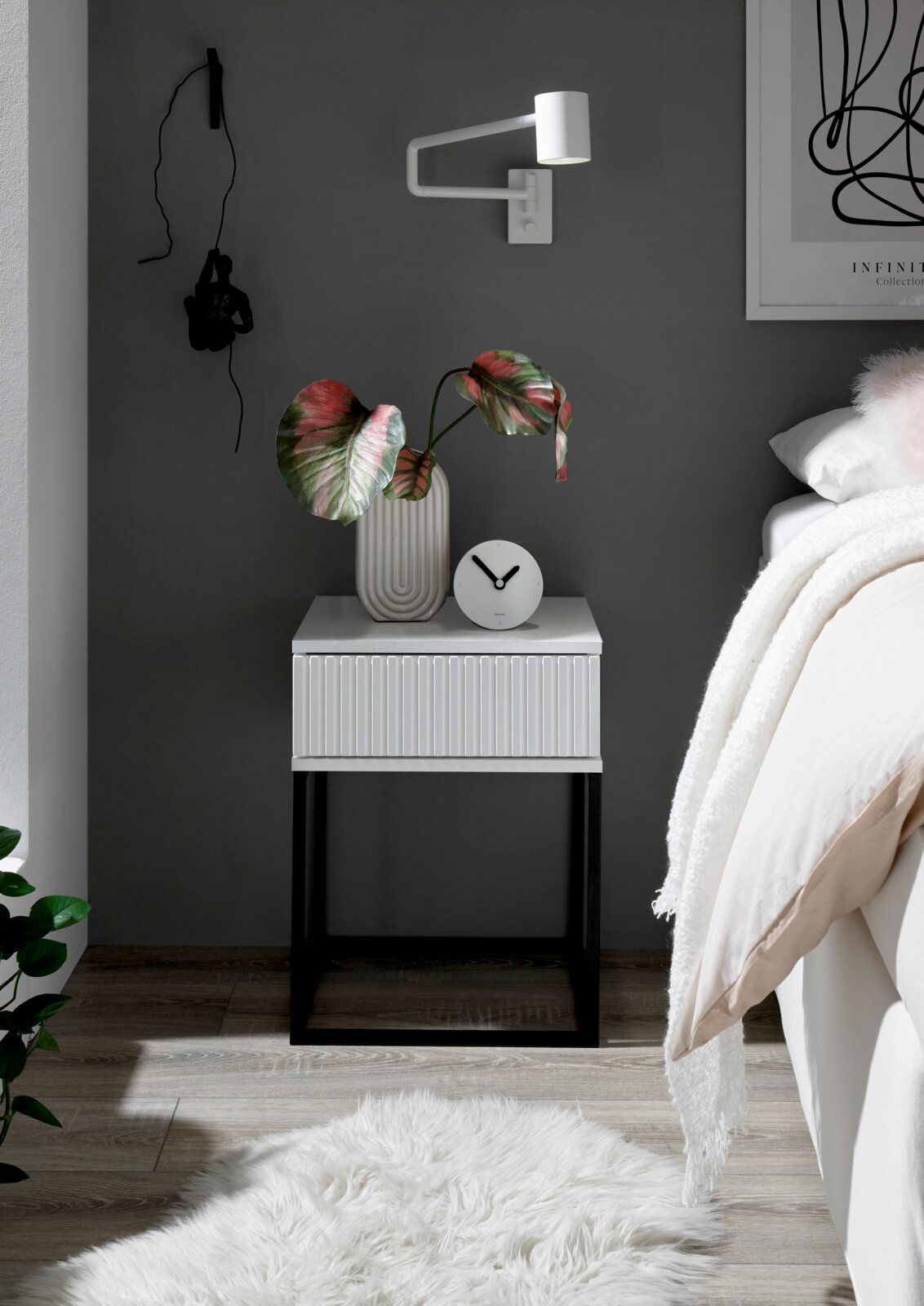 Moderne Nachtkommode mit weißer Oberfläche und schwarzen Metallbeinen, dekoriert mit einer Vase und einer Uhr, seitliche Perspektive.