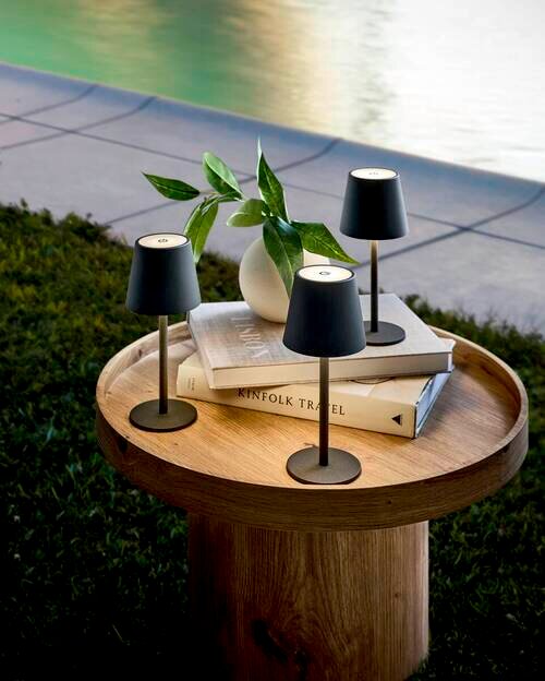 Drei moderne Akku-Tischleuchten auf einem runden Holztisch im Freien, umgeben von Büchern und einer Vase mit Pflanzen, aus der Vogelperspektive.