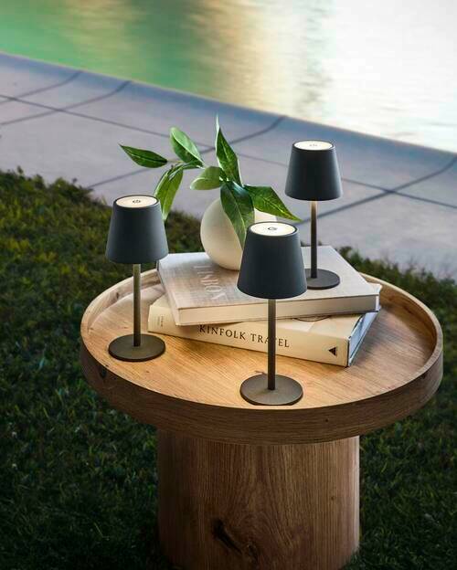 Novel Akku-Tischleuchte 3er-Set Drei moderne Akku-Tischleuchten auf einem runden Holztisch im Freien, umgeben von Büchern und einer Vase mit Pflanzen, aus der Vogelperspektive.
