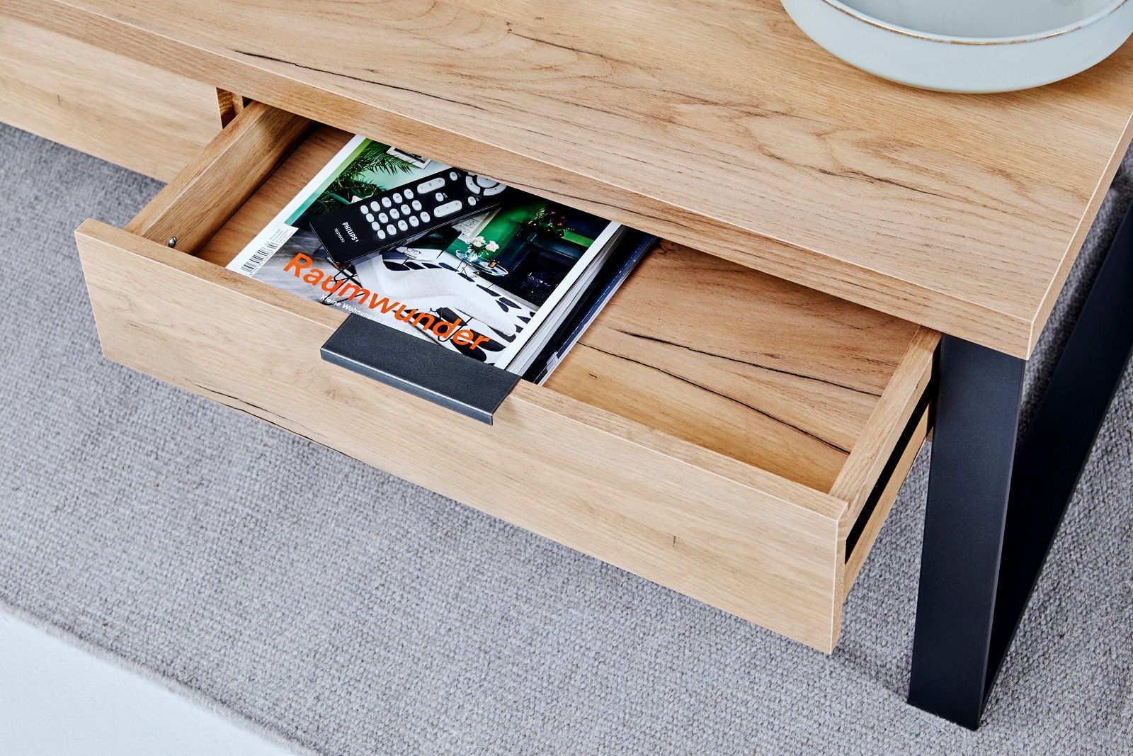 Nahaufnahme eines Couchtisches aus Holz mit schwarzem Metallgestell, einer geöffneten Schublade mit Zeitschriften und Fernbedienungen, von oben fotografiert.