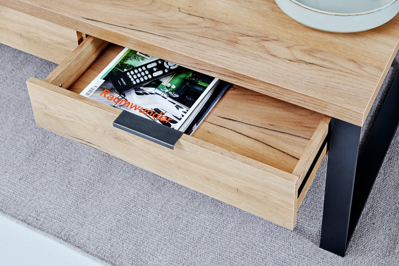 Nahaufnahme eines Couchtisches aus Holz mit schwarzem Metallgestell, einer geöffneten Schublade mit Zeitschriften und Fernbedienungen, von oben fotografiert.