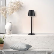 Schwarze Akku-Tischleuchte BOTERIA auf einem weißen Regal, neben einer weißen Vase mit Trockenblumen, aus seitlicher Perspektive fotografiert.