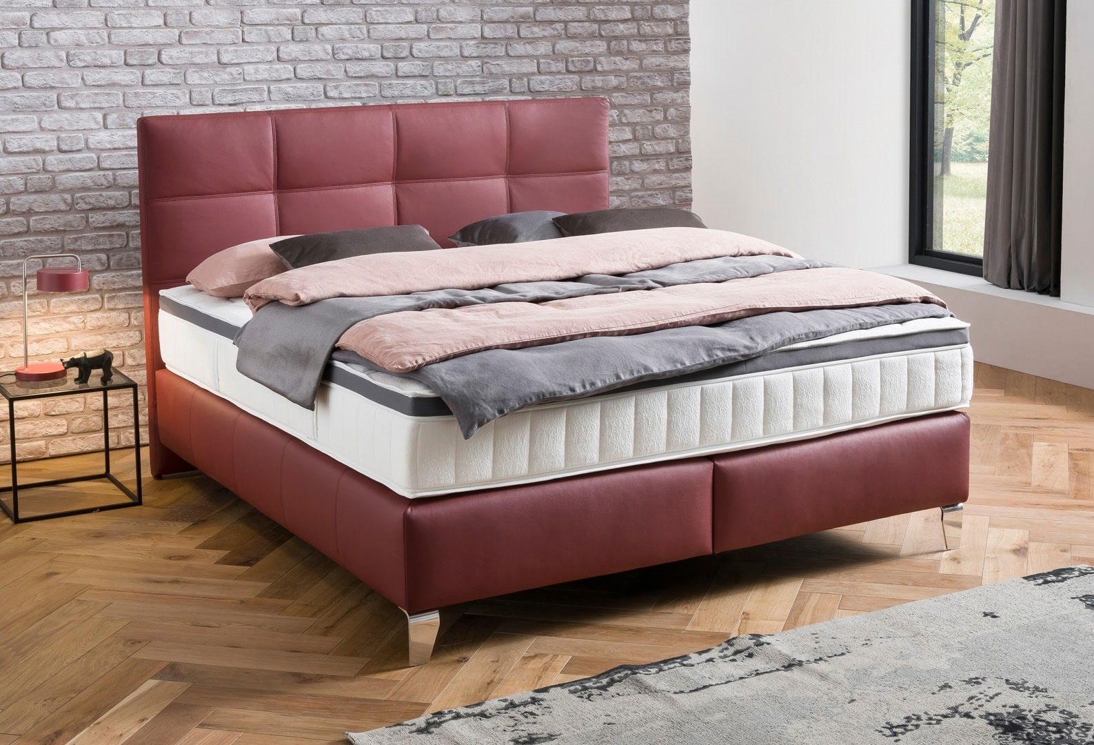 Boxspringbett mit rotem Kopfteil und Bettgestell, seitliche Perspektive, auf Parkettboden vor einer Ziegelwand.
