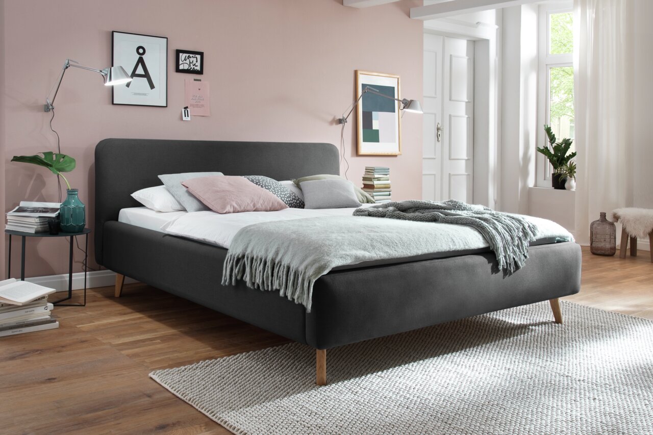 Modernes Polsterbett in dunklem Grau mit Holzfüßen, seitliche Perspektive, dekoriert mit Kissen und Decken in einem stilvoll eingerichteten Schlafzimmer.