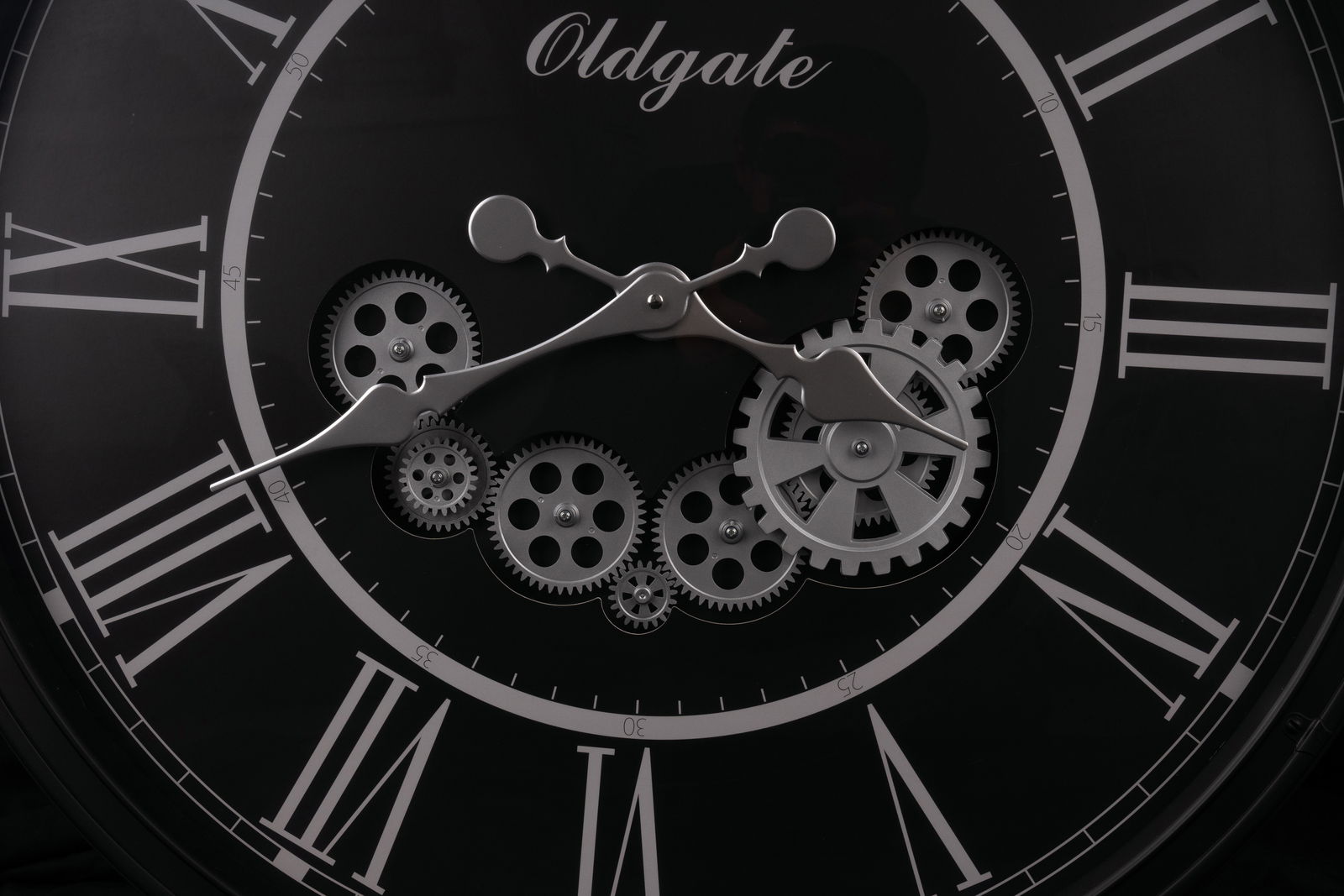 Nahaufnahme einer schwarzen Wanduhr mit römischen Ziffern und sichtbaren Zahnrädern im Inneren, frontal betrachtet.