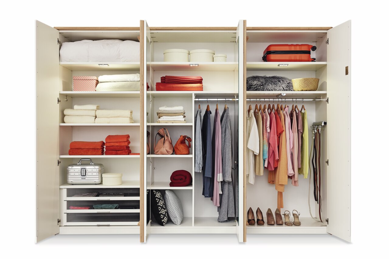 Offener 6-türiger Drehtürenschrank mit verschiedenen Fächern, Kleiderstangen und Regalen, gefüllt mit Kleidung, Handtüchern und Accessoires, aus frontaler Perspektive.