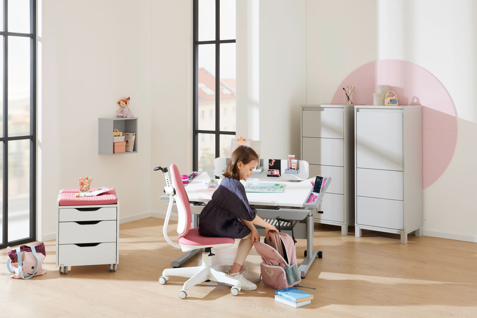 Ein moderner Jugenddrehstuhl in einem hellen, freundlichen Raum. Der Stuhl hat eine weiße Basis und eine pinke Sitzfläche. Perspektive von der Seite, mit einem Kind, das am Schreibtisch sitzt.