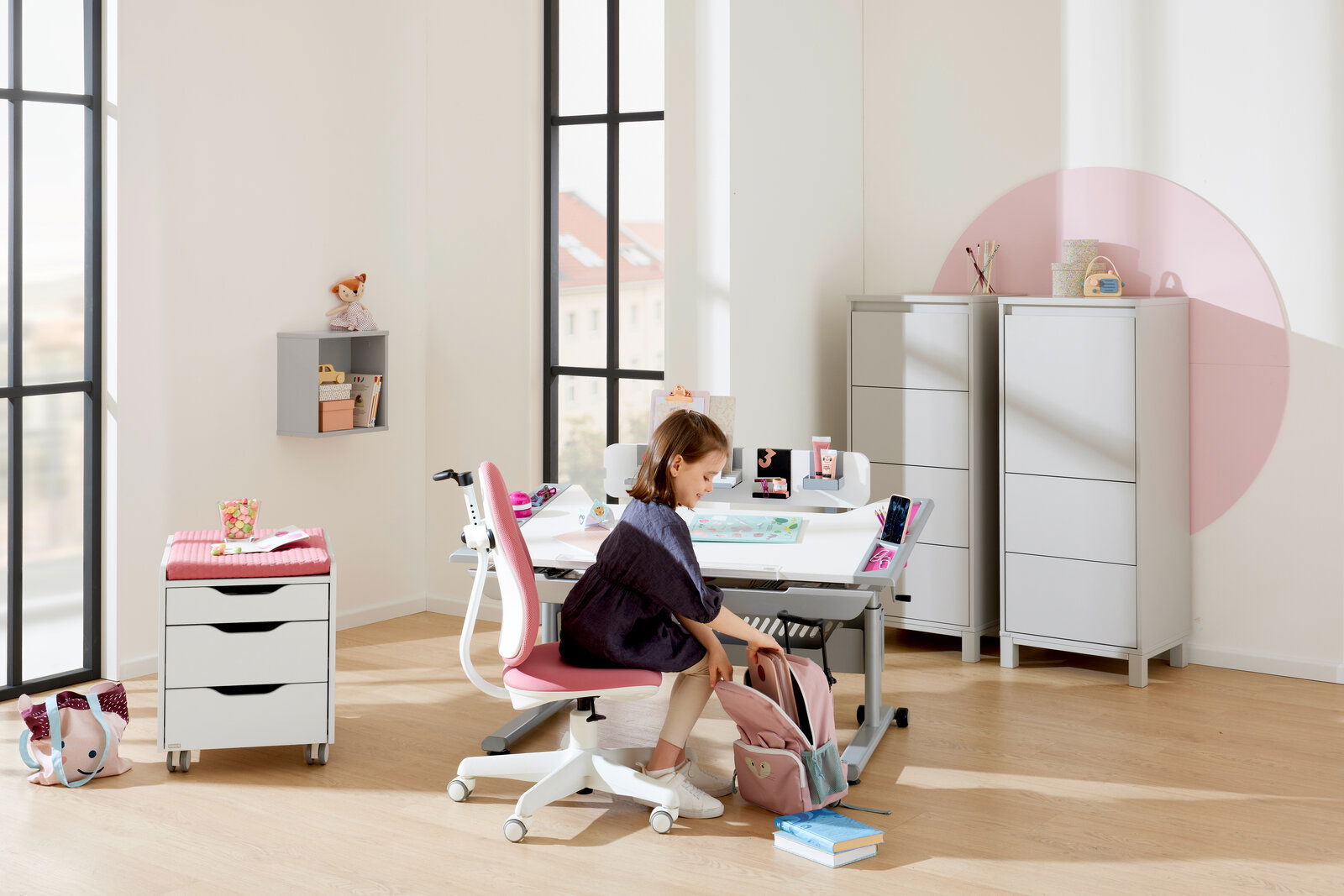 Ein moderner Jugenddrehstuhl in einem hellen, freundlichen Raum. Der Stuhl hat eine weiße Basis und eine pinke Sitzfläche. Perspektive von der Seite, mit einem Kind, das am Schreibtisch sitzt.