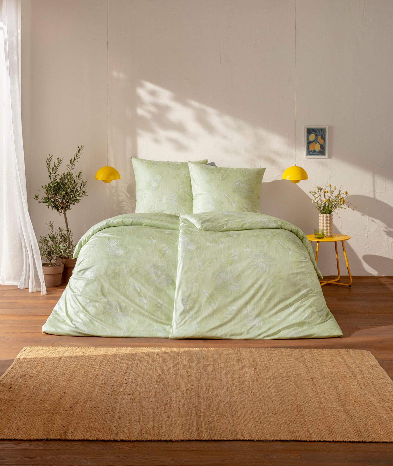 Hellgrüne Bettwäsche mit dezentem floralem Muster auf einem Bett im sonnigen Schlafzimmer, frontal aus leicht erhöhter Perspektive fotografiert gelbe Pendelleuchten, gelber Beistelltisch und Pflanzen im Hintergrund.