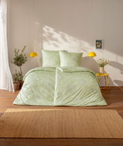Hellgrüne Bettwäsche mit dezentem floralem Muster auf einem Bett im sonnigen Schlafzimmer, frontal aus leicht erhöhter Perspektive fotografiert; gelbe Pendelleuchten, gelber Beistelltisch und Pflanzen im Hintergrund.