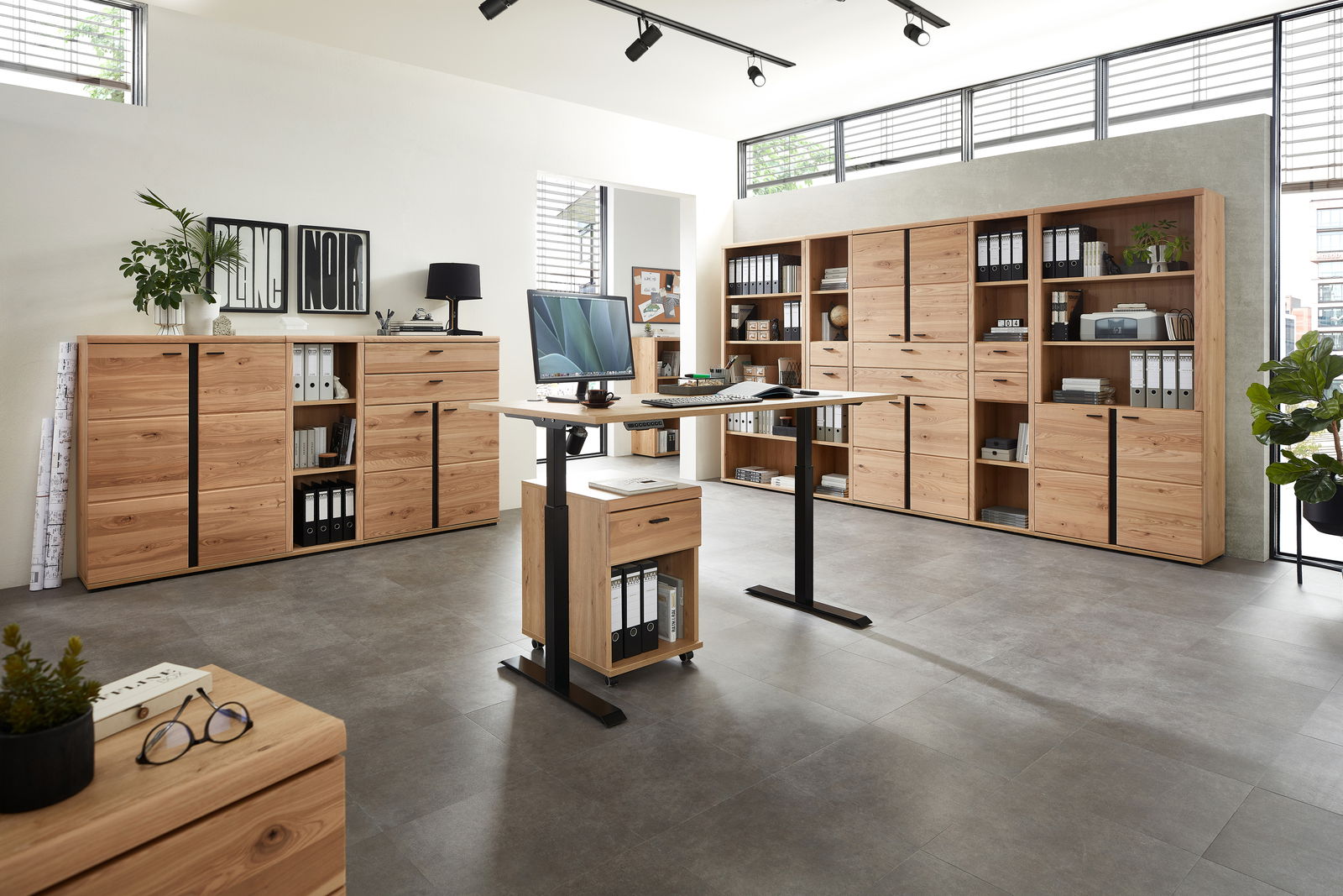 Moderne Büromöbel in einem hellen Raum mit großen Fenstern, bestehend aus einem höhenverstellbaren Schreibtisch und mehreren Aktenschränken aus Holz. Perspektive von vorne.