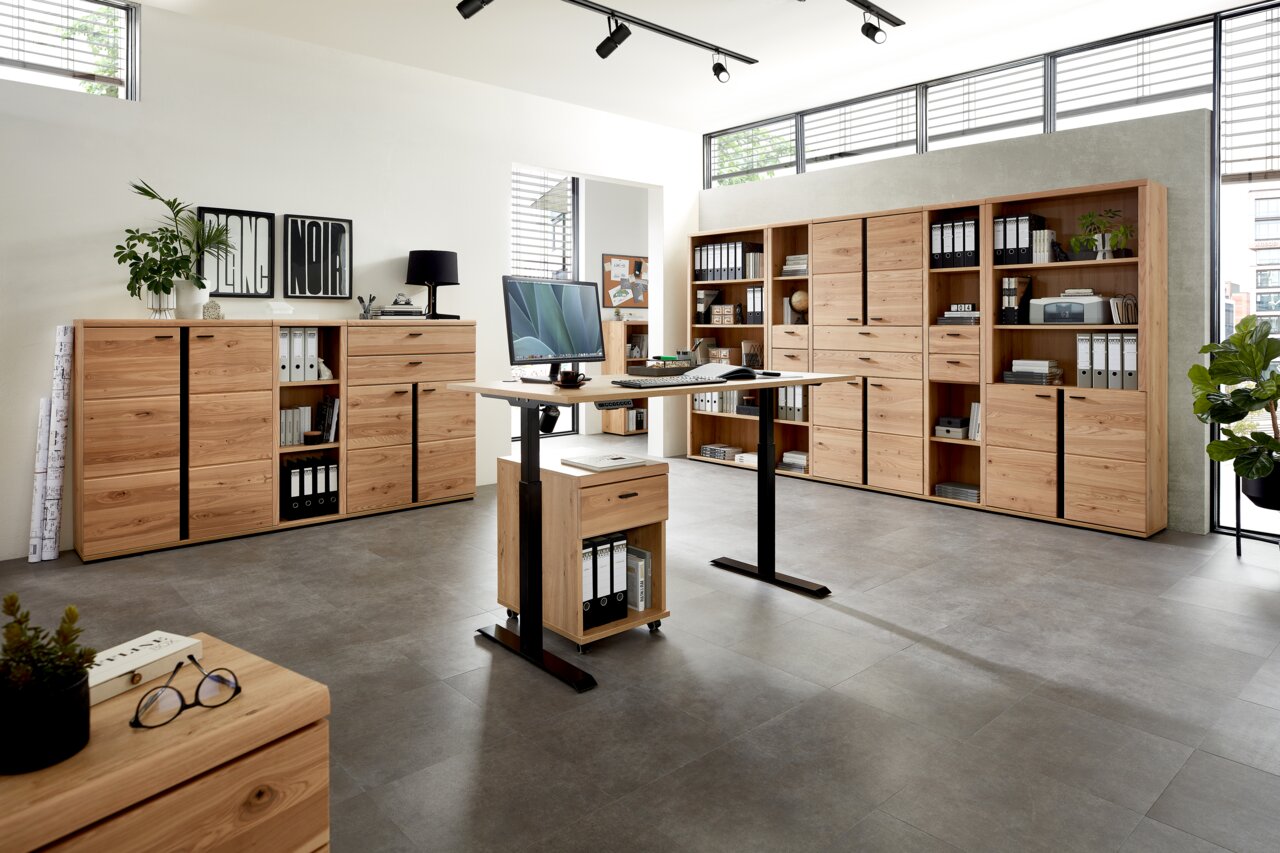 Moderne Büromöbel in einem hellen Raum mit großen Fenstern, bestehend aus einem höhenverstellbaren Schreibtisch und mehreren Aktenschränken aus Holz. Perspektive von vorne.