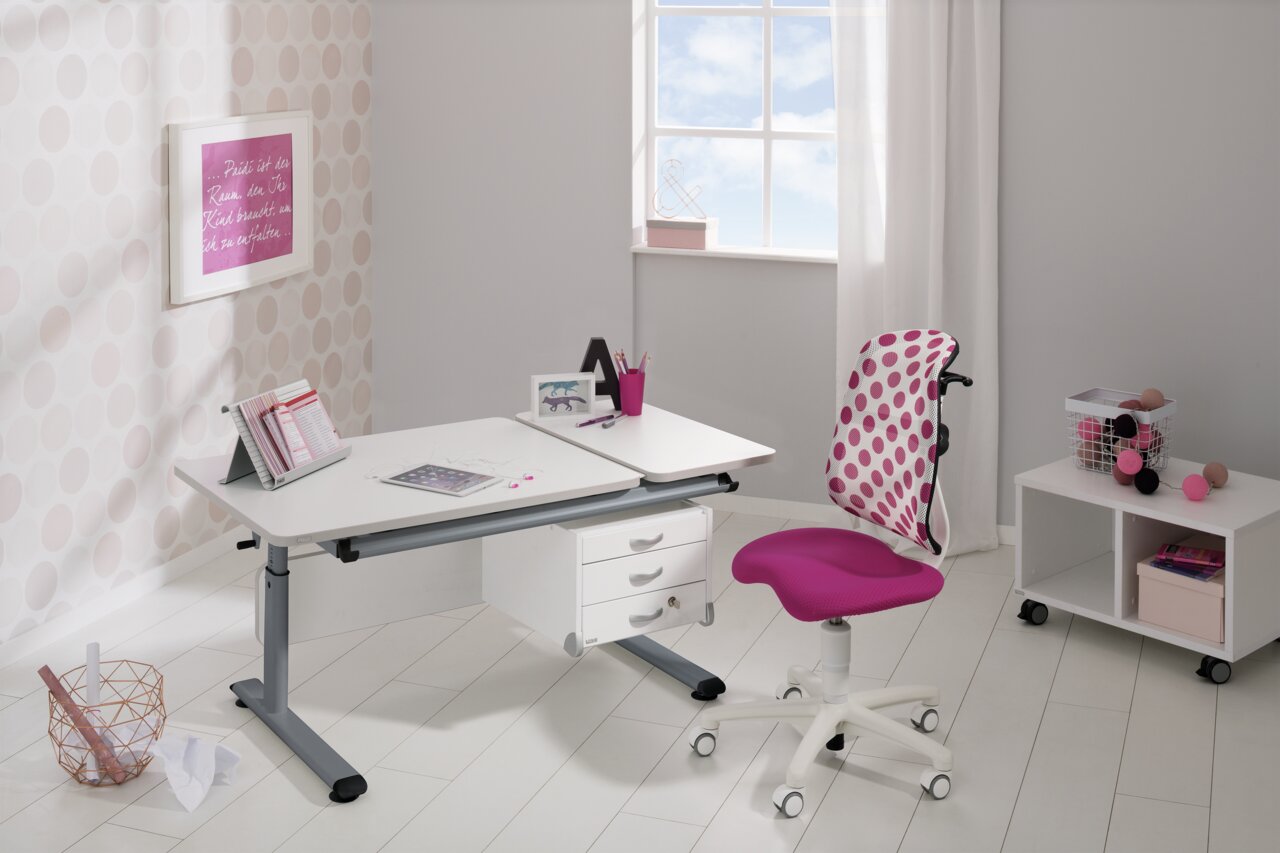 Moderner Schreibtisch in einem hellen Raum mit einem großen Fenster im Hintergrund. Der Schreibtisch hat eine weiße Oberfläche und graue Beine, ausgestattet mit Schubladen. Ein pink gepunkteter Stuhl steht davor. Perspektive: seitliche Ansicht.