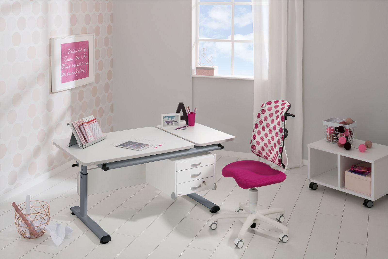 Moderner Schreibtisch in einem hellen Raum mit einem großen Fenster im Hintergrund. Der Schreibtisch hat eine weiße Oberfläche und graue Beine, ausgestattet mit Schubladen. Ein pink gepunkteter Stuhl steht davor. Perspektive: seitliche Ansicht.