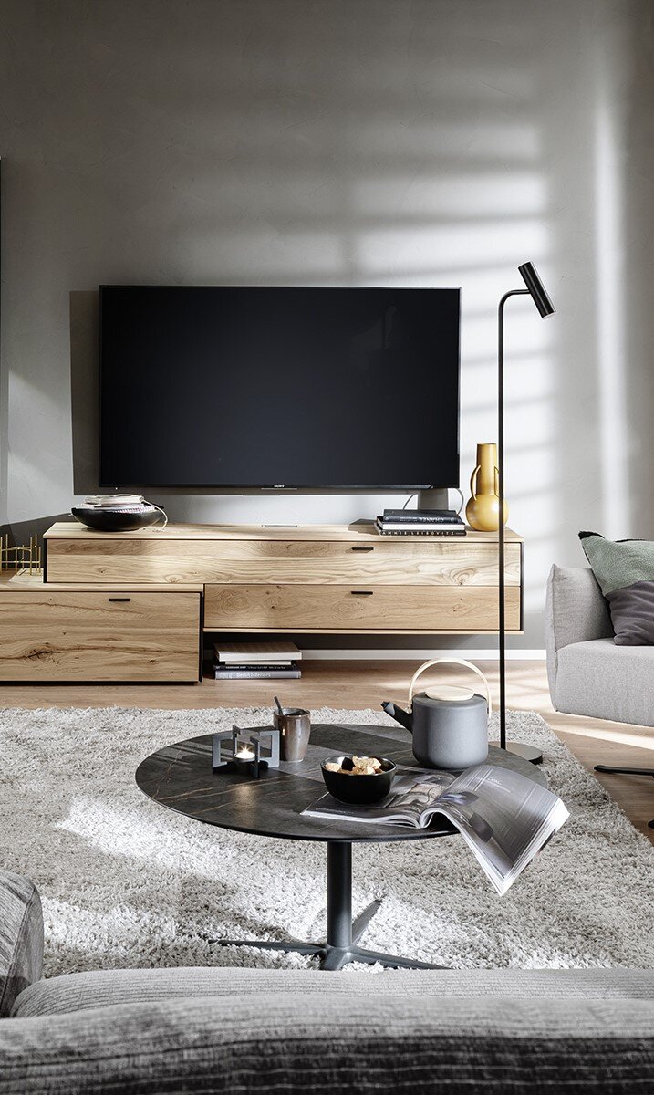 Moderne wandcombinatie ANDRA in licht hout met ingebouwde tv, gezien vanaf de voorkant. Stijlvolle decoraties en een gezellige zithoek op de voorgrond.