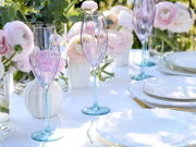 Zart violett getöntes Champagnerglas mit türkisfarbenem Stiel auf festlich gedecktem Tisch mit pastellfarbenen Blumen und weißen Tellern, fotografiert aus leicht schräger Seitenperspektive auf Tischhöhe.