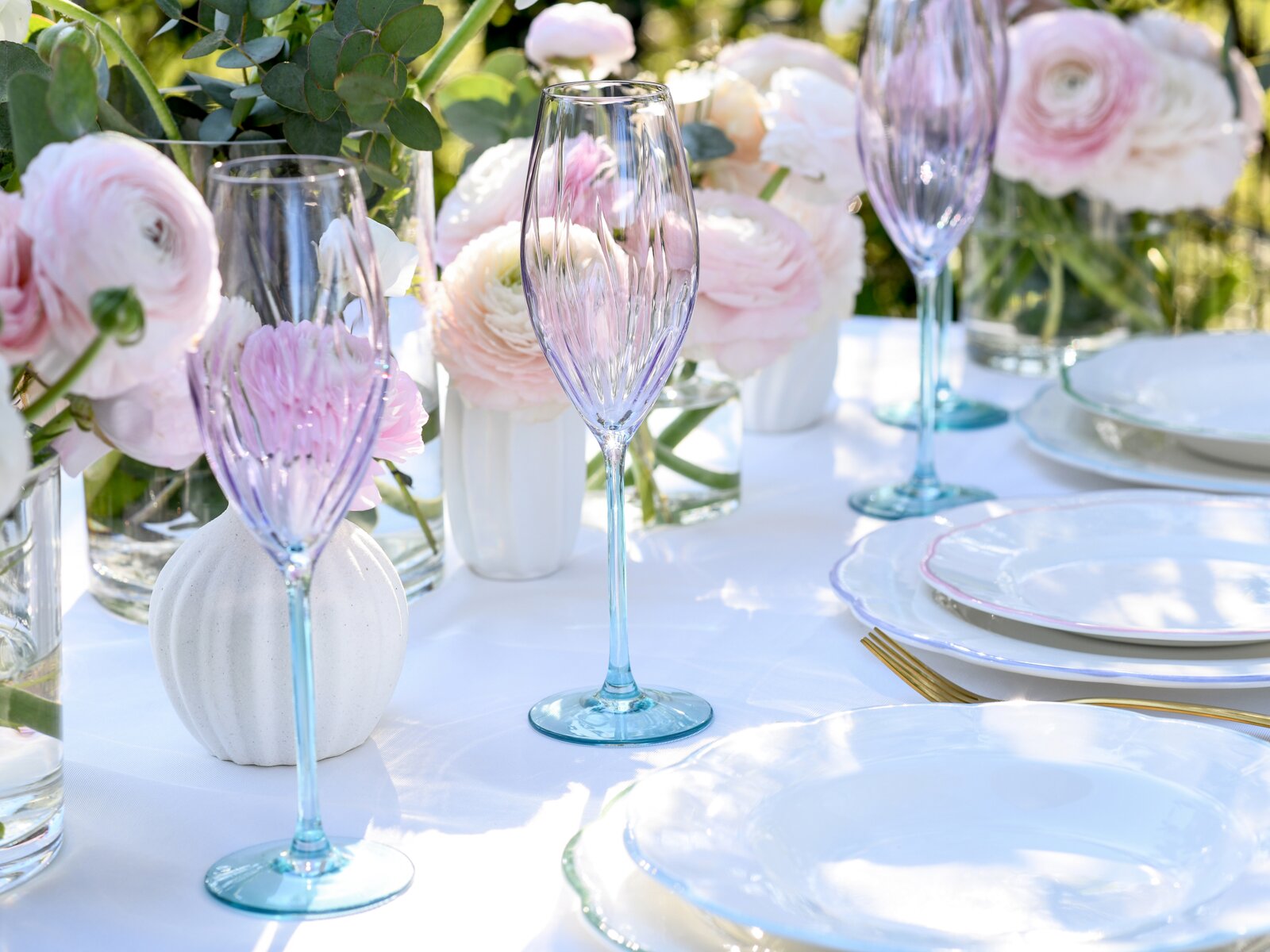 Zart violett getöntes Champagnerglas mit türkisfarbenem Stiel auf festlich gedecktem Tisch mit pastellfarbenen Blumen und weißen Tellern, fotografiert aus leicht schräger Seitenperspektive auf Tischhöhe.