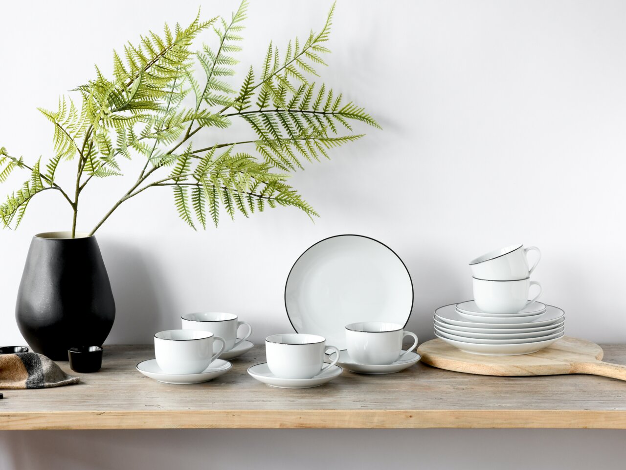 Kaffeeservice 18-teilig Luna Black L, bestehend aus Tassen, Untertassen und Tellern mit schwarzem Rand, auf einem Holztisch mit schwarzer Vase und grünen Zweigen, seitliche Perspektive.