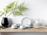 Kaffeeservice 18-teilig Luna Black L, bestehend aus Tassen, Untertassen und Tellern mit schwarzem Rand, auf einem Holztisch mit schwarzer Vase und grünen Zweigen, seitliche Perspektive.