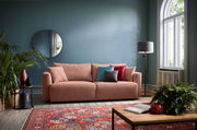 Ein modernes Funktionssofa in einem gemütlichen Wohnzimmer, frontal fotografiert. Das Sofa ist in einem warmen Rosaton gehalten und mit bunten Kissen dekoriert. Im Hintergrund sind eine blaue Wand, ein runder Spiegel und eine Stehlampe zu sehen.