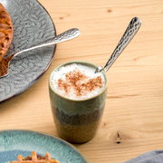 Een gebogen latte macchiato-lepel van roestvrij staal in een kopje met melkschuim, gefotografeerd van bovenaf.