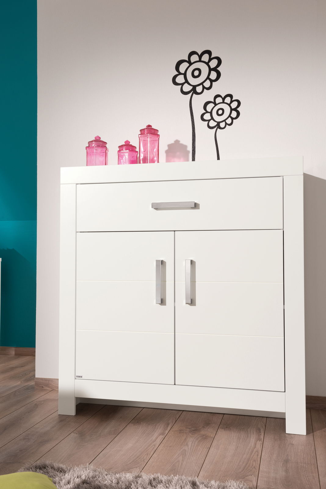 Weißes Highboard mit zwei Türen und einer Schublade, dekoriert mit rosa Gläsern, aus frontaler Perspektive fotografiert.