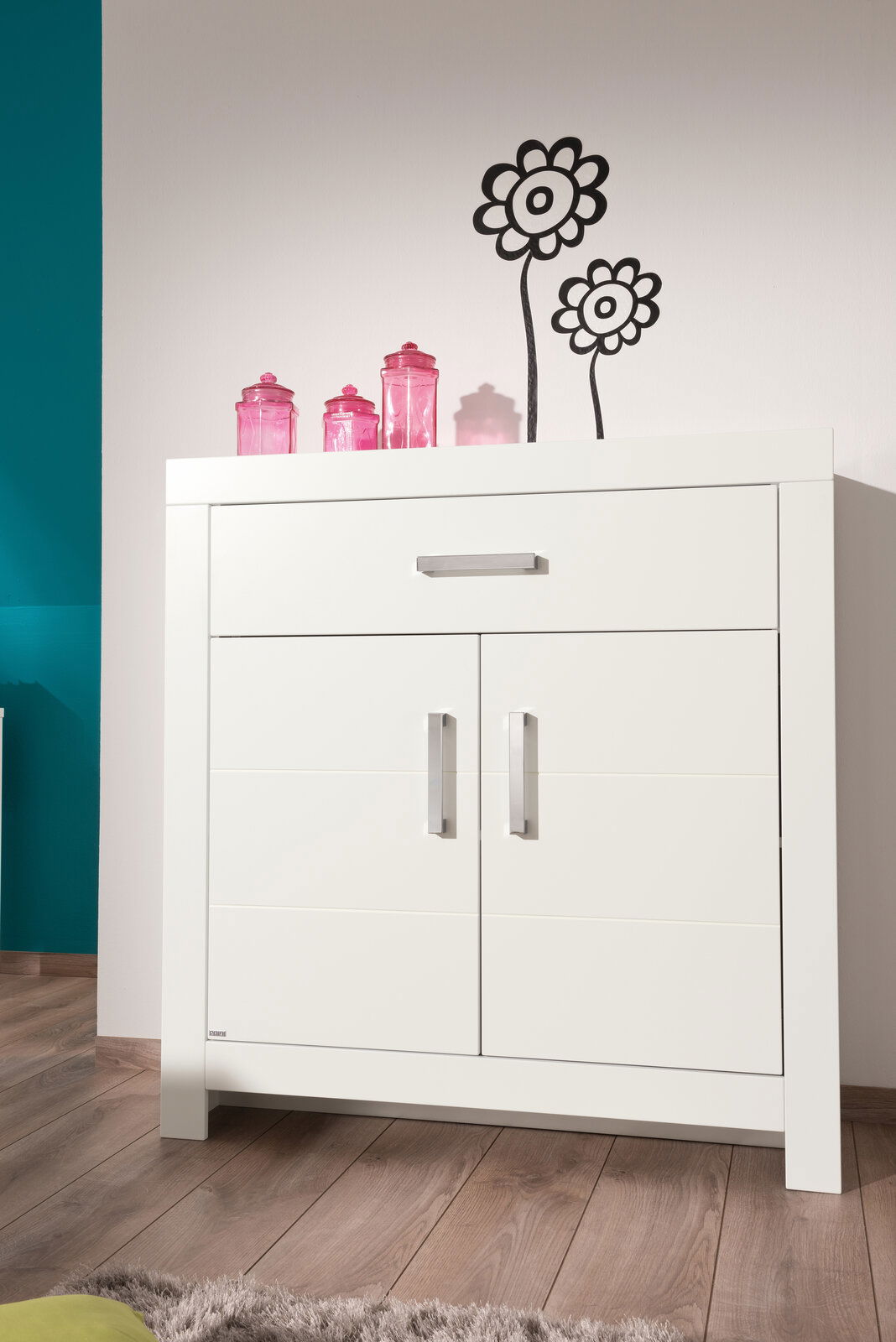 Weißes Highboard mit zwei Türen und einer Schublade, dekoriert mit rosa Gläsern, aus frontaler Perspektive fotografiert.