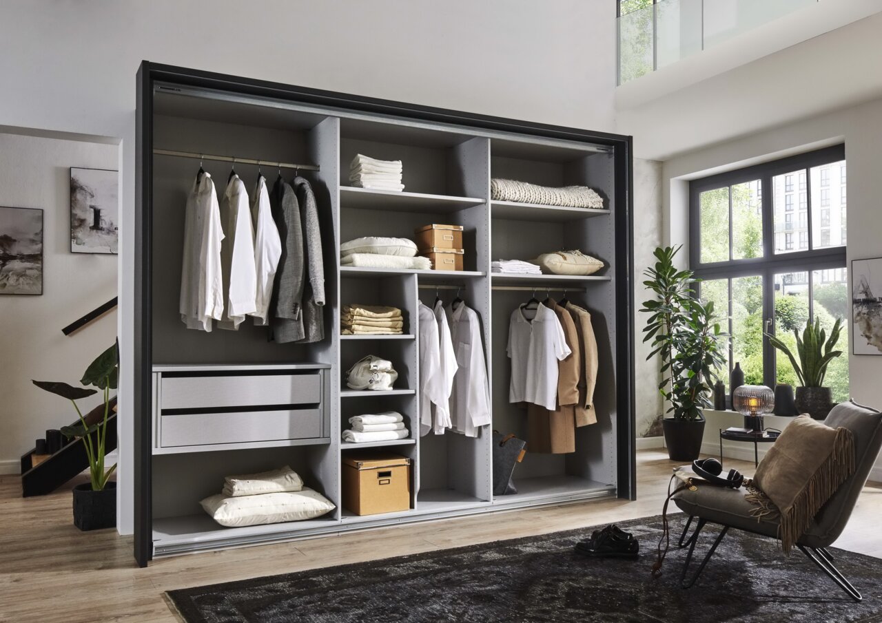 Open kledingkast met grijze binnenbekleding, gevuld met kleding en accessoires, in een moderne woonkamer met planten en grote ramen, gezien van voren.