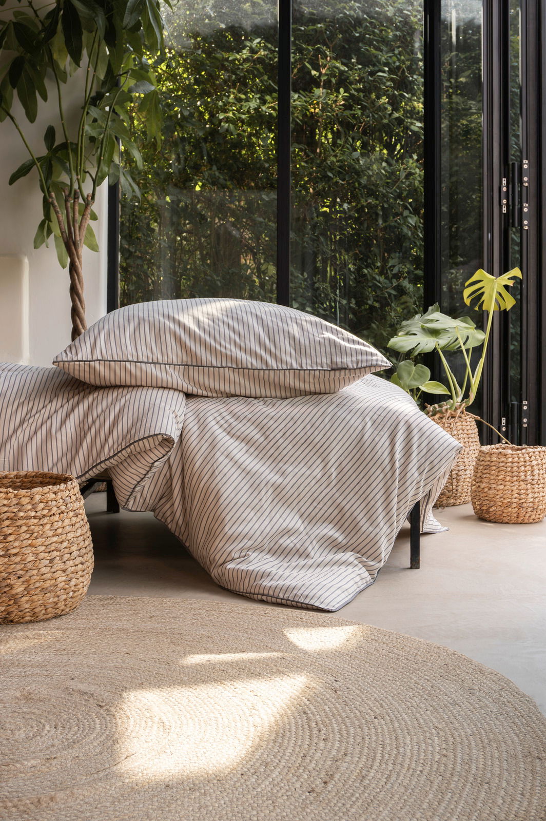 Bettwäsche-Set mit feinen Streifen in einem modernen, hellen Raum mit großen Fenstern und Pflanzen, aus der Seitenperspektive fotografiert.