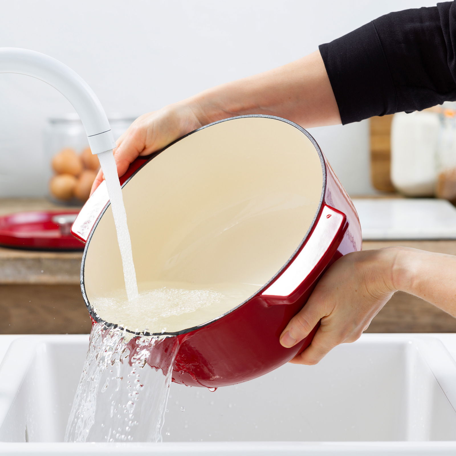 Roter ovaler Gusseisen-Bräter mit cremefarbener Innenbeschichtung, der in einem Waschbecken unter fließendem Wasser gereinigt wird. Perspektive von der Seite.