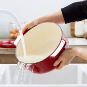 Roter ovaler Gusseisen-Bräter mit cremefarbener Innenbeschichtung, der in einem Waschbecken unter fließendem Wasser gereinigt wird. Perspektive von der Seite.