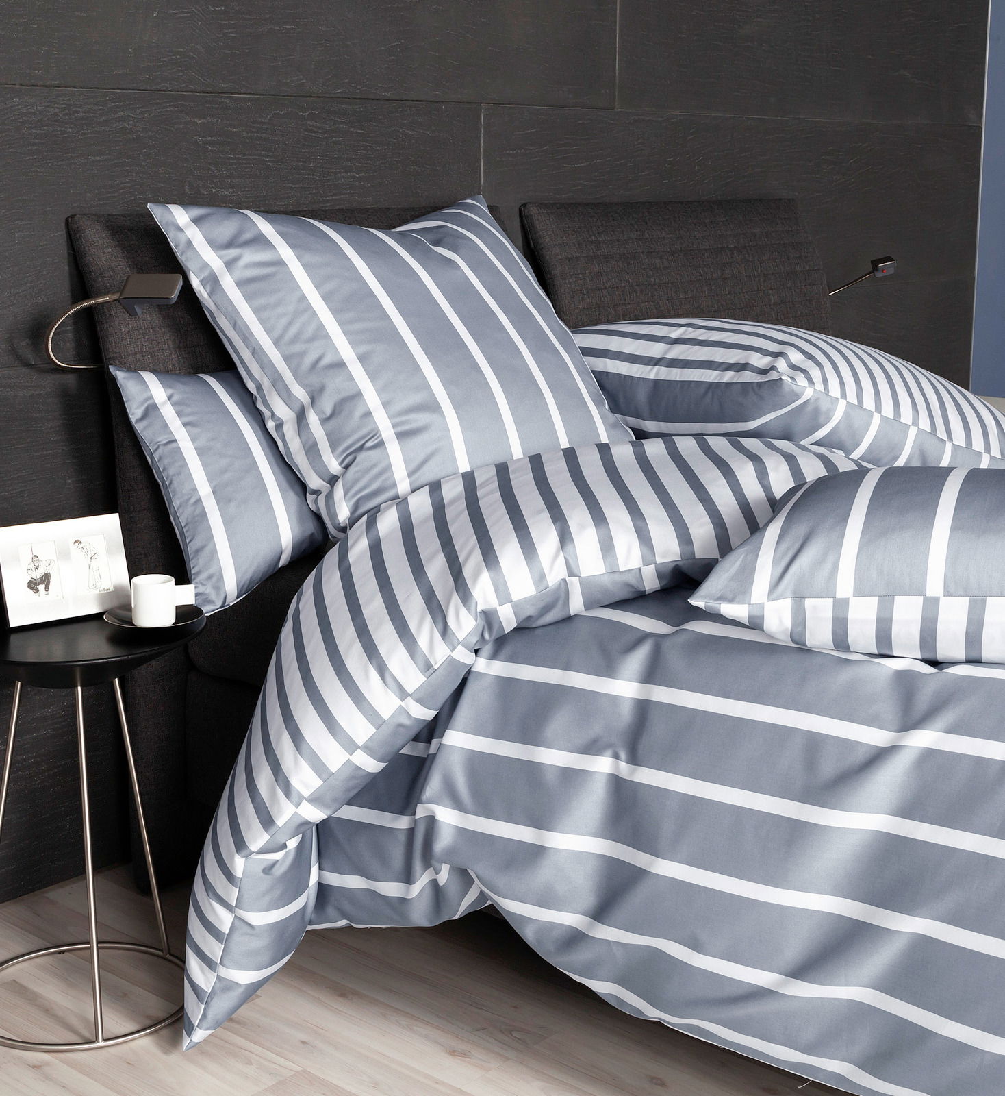 Bettwäsche-Set in Grau mit weißen Streifen, auf einem Bett mit dunklem Kopfteil, seitliche Perspektive
