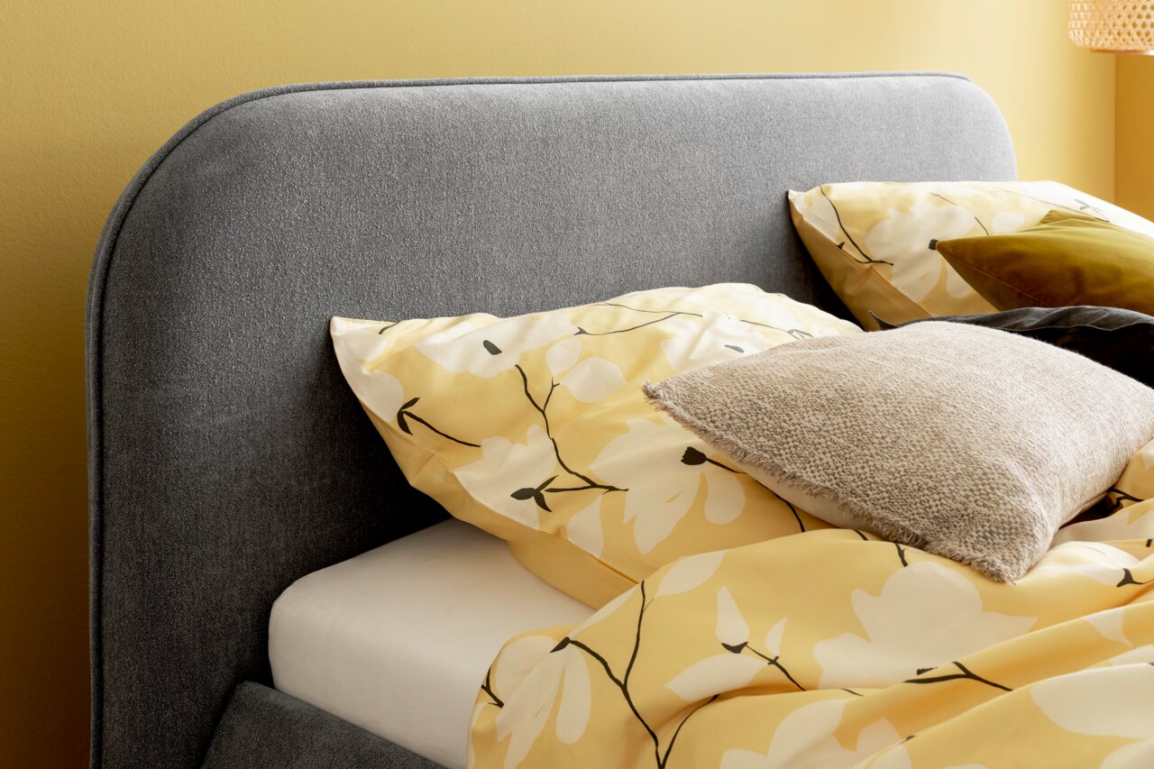 Graues Polsterbett mit gelber Blumenbettwäsche und Kissen, seitliche Perspektive