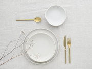 Minimalistisches weißes Geschirr-Set mit zwei Tellern und einer Schale sowie goldfarbenem Besteck auf hellem Leinen, Draufsicht (Flatlay)