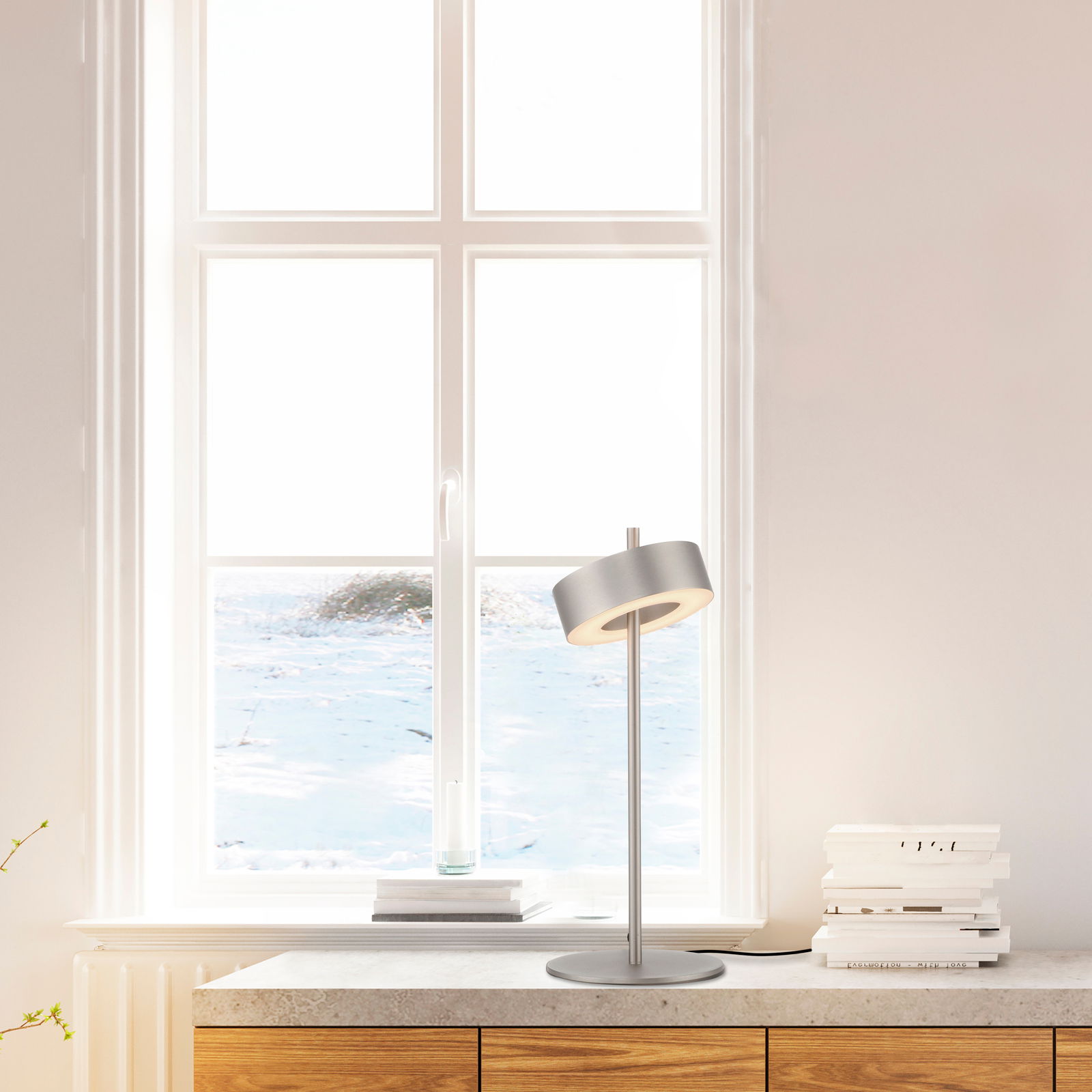 Moderne Tischleuchte Q-ROTATE in silberner Farbe, seitliche Perspektive, auf einem Tisch vor einem Fenster platziert.